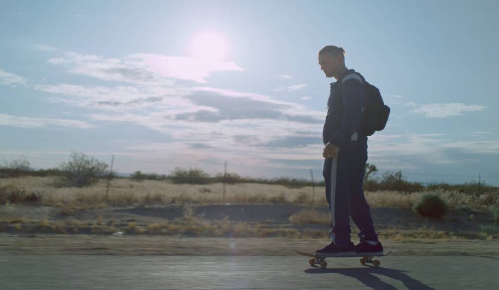 Garoto andando de skate com mochila nas costas e sol ao fundo